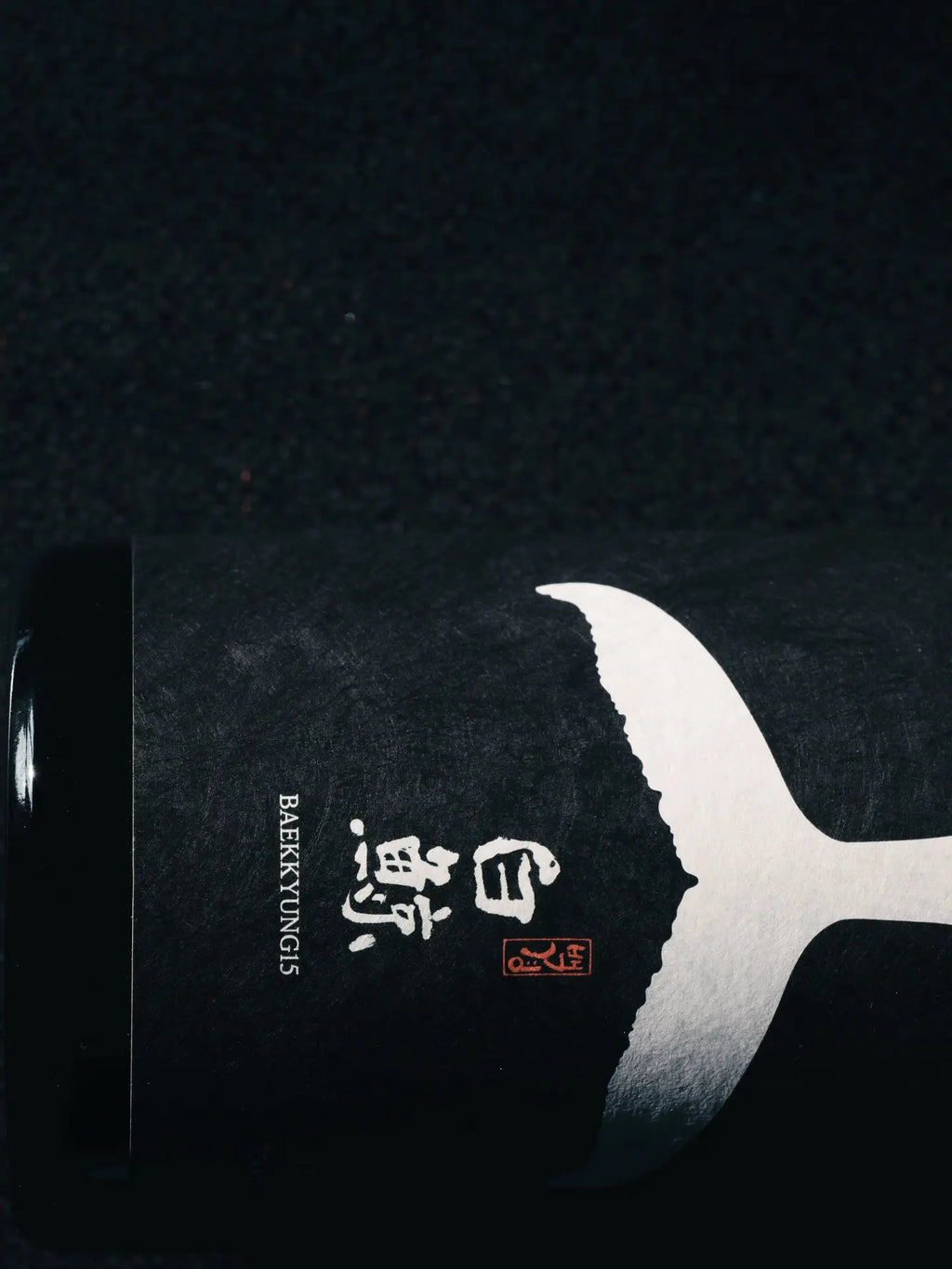 A closeup of a black hanji paper wine bottle label against a black background. The label has a white whale tail logo graphic, hanja characters, and the word BAEKKYUNG15. 