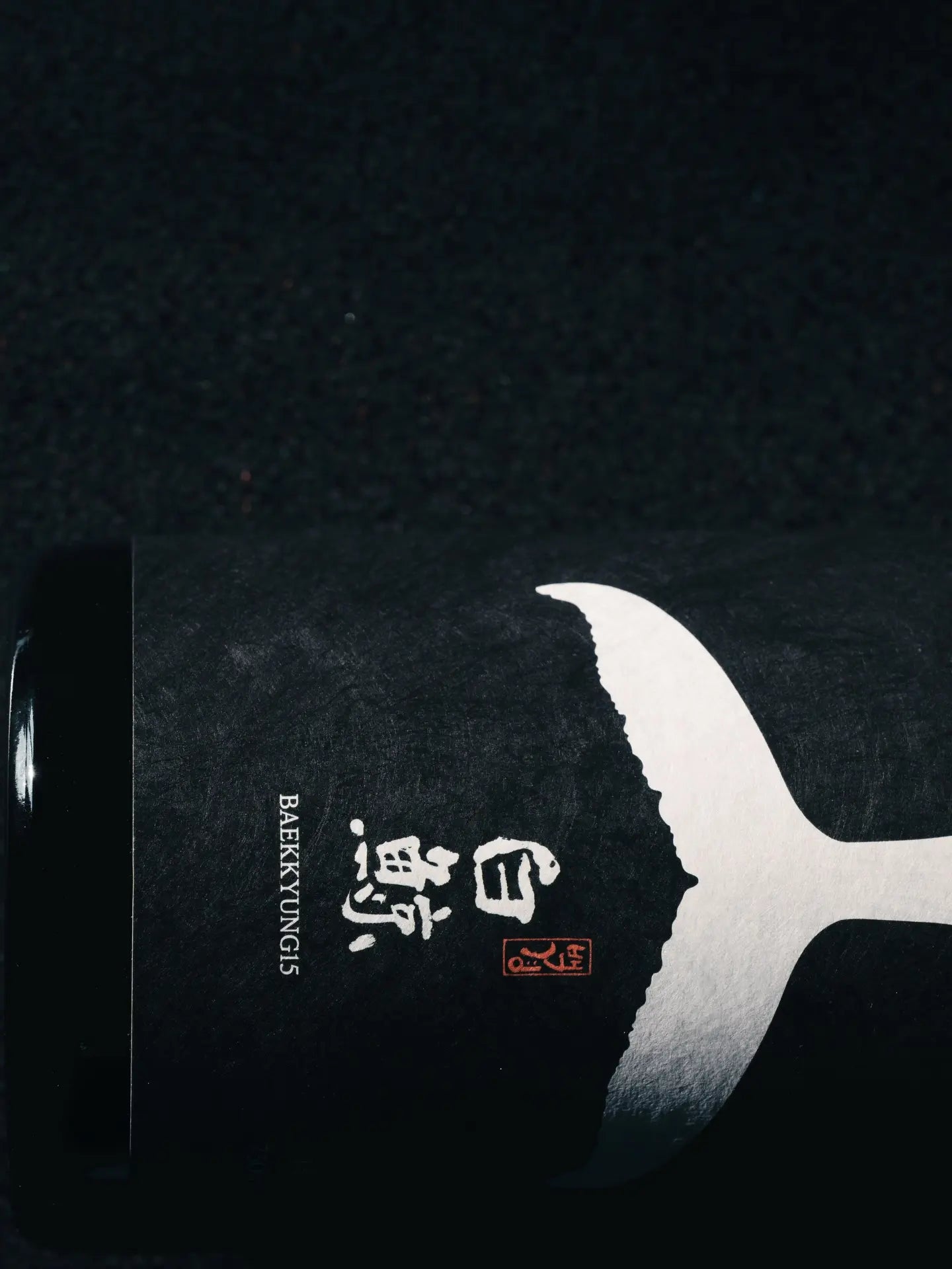 A closeup of a black hanji paper wine bottle label against a black background. The label has a white whale tail logo graphic, hanja characters, and the word BAEKKYUNG15. 