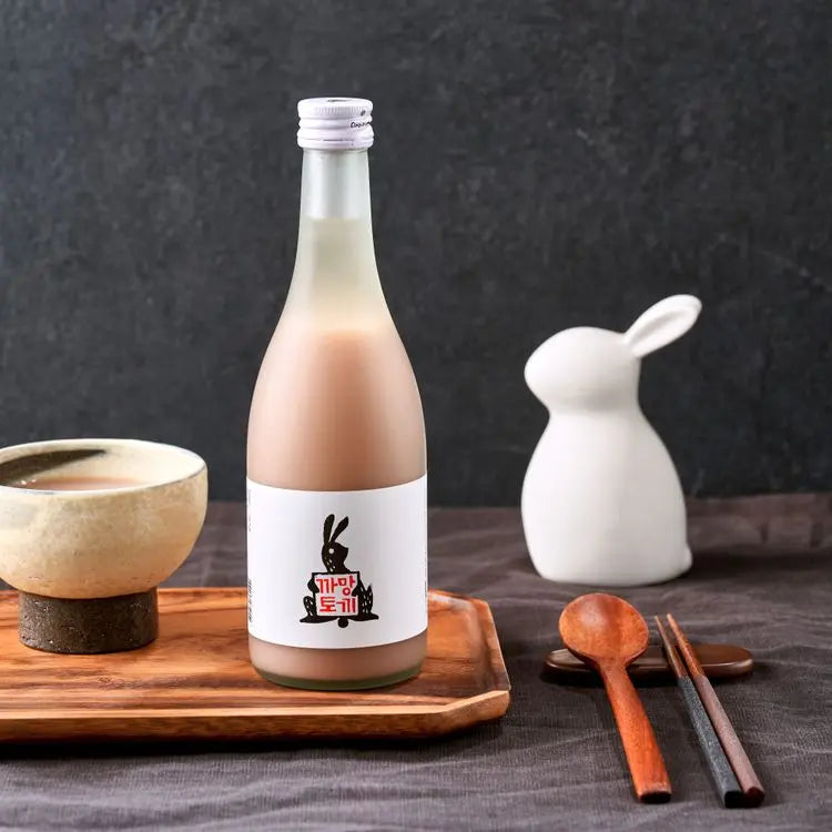 A bottle of Korean rice wine with a white label featuring a black rabbit illustration and red text, set on a wooden tray next to a ceramic bowl, a white rabbit-shaped vessel, and wooden utensils against a dark gray background.

Black Rabbit Makgeolli - Sulsaem - cultsool