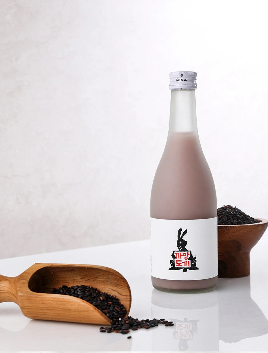 A clear glass bottle filled with opaque pink-brown makgeolli stands on a white surface against a light gray background.  In the foreground, a wooden scoop filled with black rice.

Black Rabbit Makgeolli - Sulsaem - cultsool