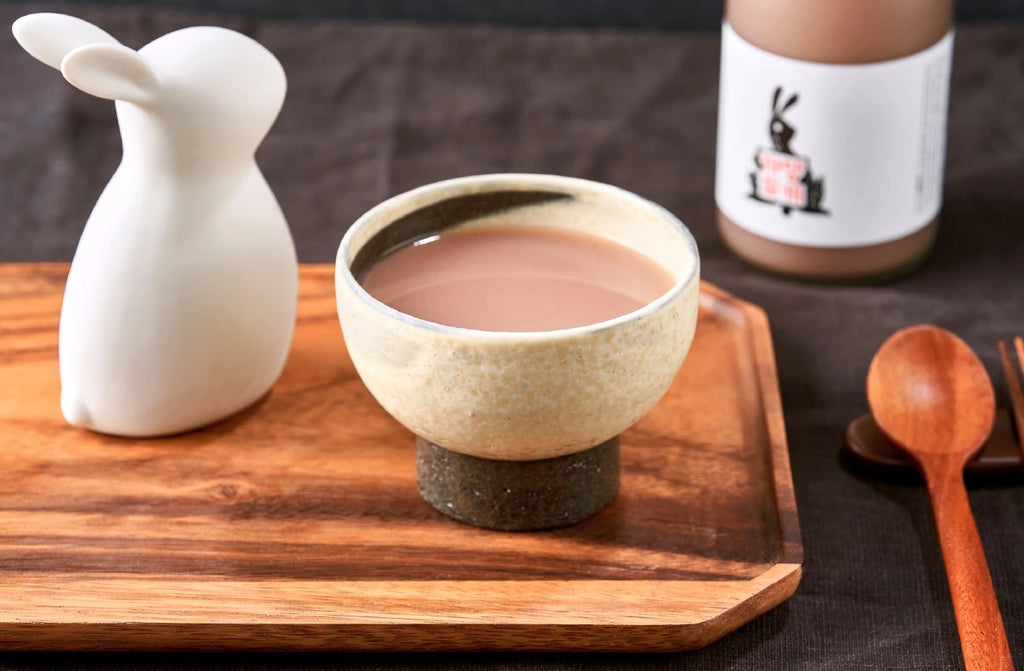 A white ceramic rabbit-shaped object and a speckled beige bowl filled with a light pink-brown rice wine, arranged on a wooden tray with a wooden spoon, set against a dark textured background. A bottle of Black Rabbit Makgeolli is visible in the background.

Black Rabbit Makgeolli - Sulsaem - cultsool