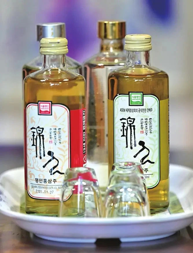Two bottles of Korean rice wine with clear glass bodies and pale yellow liquid, displayed on a white plate with small clear drinking glasses in front. The background is softly blurred with purple and blue tones.

Geumsan Ginseng Wine - Geumsan Insamju - cultsool