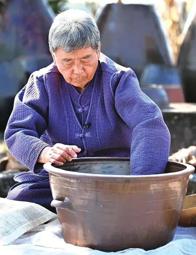 An older man (Korean Food Grandmaster) Kim Chang-soo in a textured purple traditional robe sits outdoors, dipping his hands into a large brown ceramic pot while making ginseng liquor.

Geumsan Ginseng Wine - Geumsan Insamju - cultsool