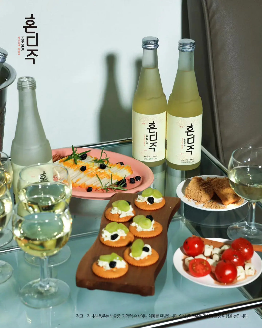 Two bottles of Korean citrus wine (Hondiju) on a glass table alongside various appetizers, wine glasses, and a glass of white wine, set against a neutral indoor background.

Hondiju - Citrus Brewery - cult