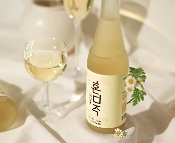 A bottle of Korean fruit win with a pale yellow liquid and a cream-colored label with black Korean text, styled on a white fabric surface with a filled wine glass, another blurred bottle, and small white flowers.
Hondiju - Citrus Brewery - cult