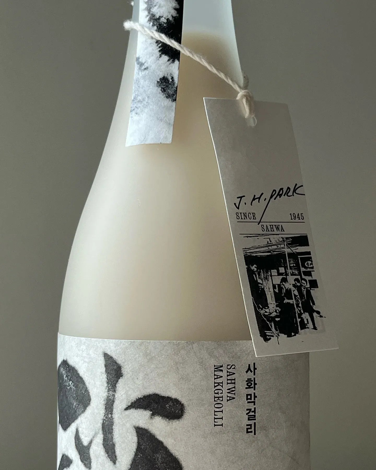 A close-up of a Korean rice wine bottle with a frosted off-white body, black ink-style label graphics, and a hanging paper tag reading J.H. Park

Sahwa Makgeolli - Good Tomorrow Brewery - cultsool
