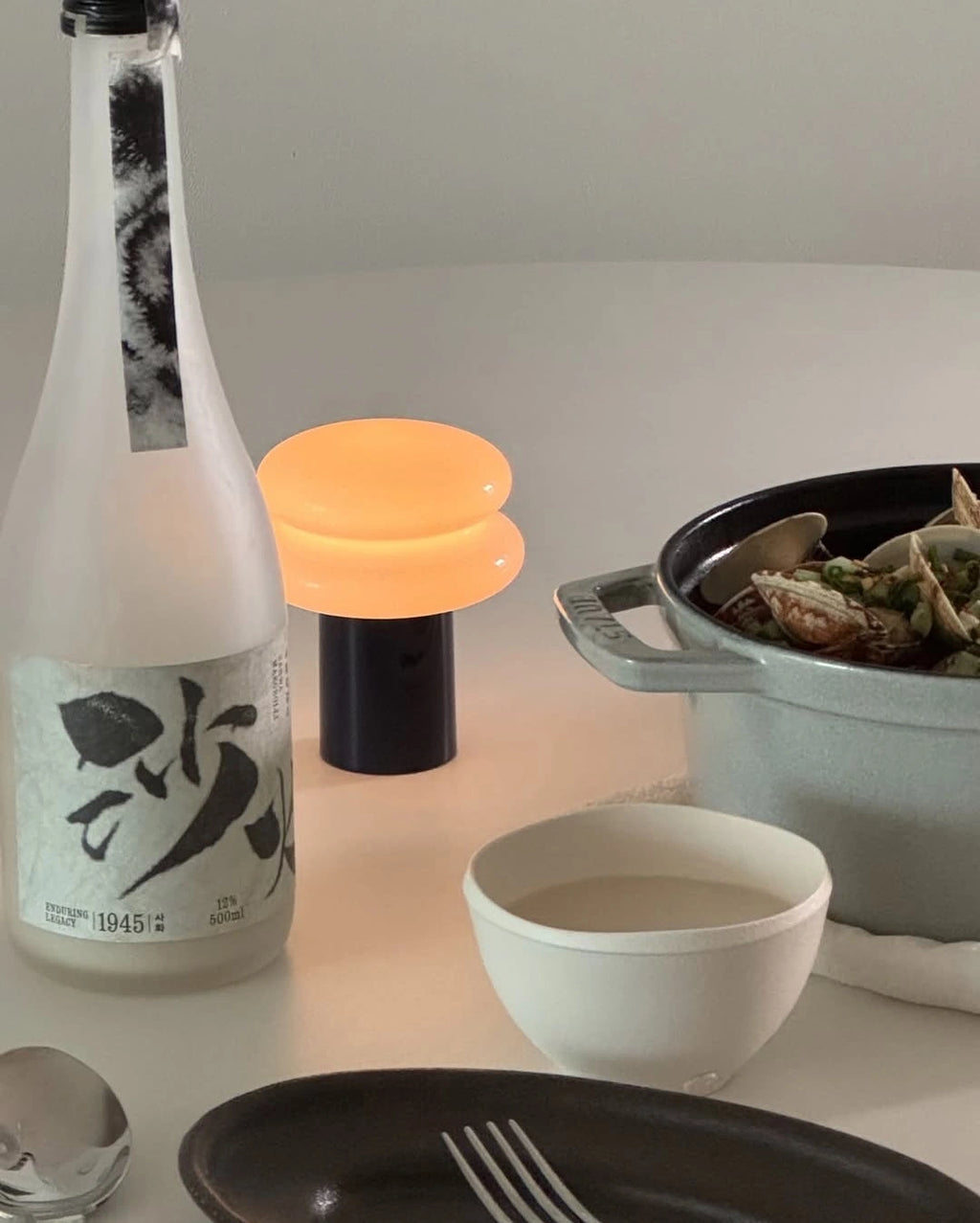 A styled tabletop scene with a frosted glass rice wine bottle, a glowing black-and-cream table lamp, a bowl of clams, and ceramic dishes on a light surface.


Sahwa Makgeolli - Good Tomorrow Brewery - cultsool