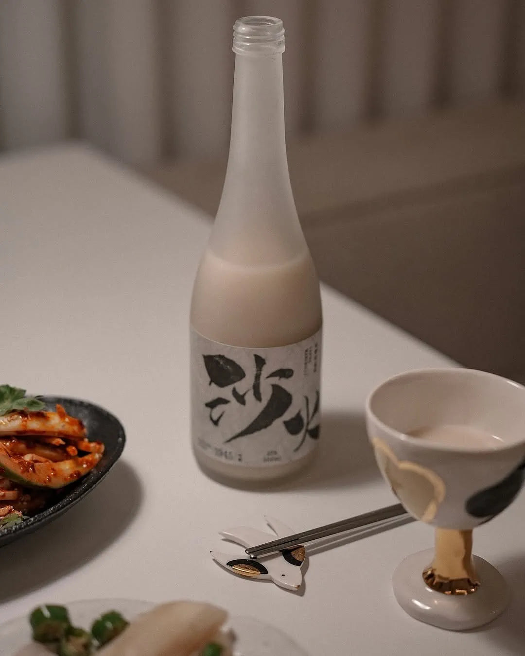 A frosted glass bottle of Korean rice wine with a white label featuring black brushstroke characters, set on a white table next to a small bowl of food, chopsticks, and a ceramic sake cup.


Sahwa Makgeolli - Good Tomorrow Brewery - cultsool