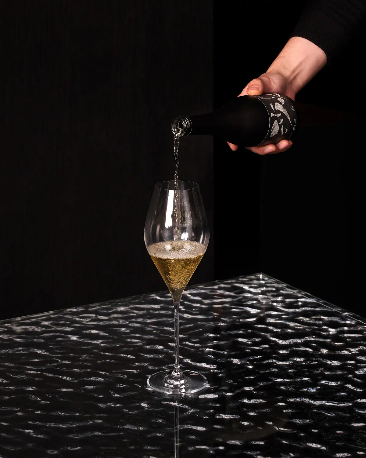 A hand pours a golden beverage from a black bottle into a clear stemmed glass on a reflective black surface against a dark background. Sahwa Yakju - Good Tomorrow Brewery - cult