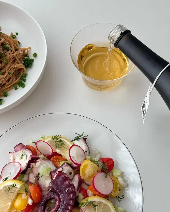 A bottle of yakju (clear Korean rice wine) is pouring into a clear glass, with a plate of octopus salad and another dish of pasta partially visible on a white tabletop.  Sahwa Yakju - Good Tomorrow Brewery - cult