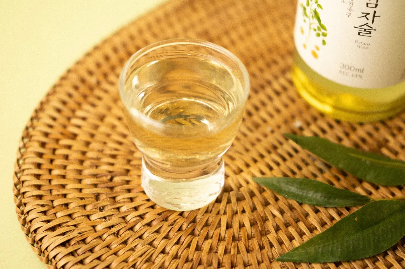 A small clear glass filled with pale golden liquid sits on a round woven straw mat, with part of a labeled bottle and green leaves visible in the background.

Seoju Gamjasool yakju - Odae Seoju - cult 