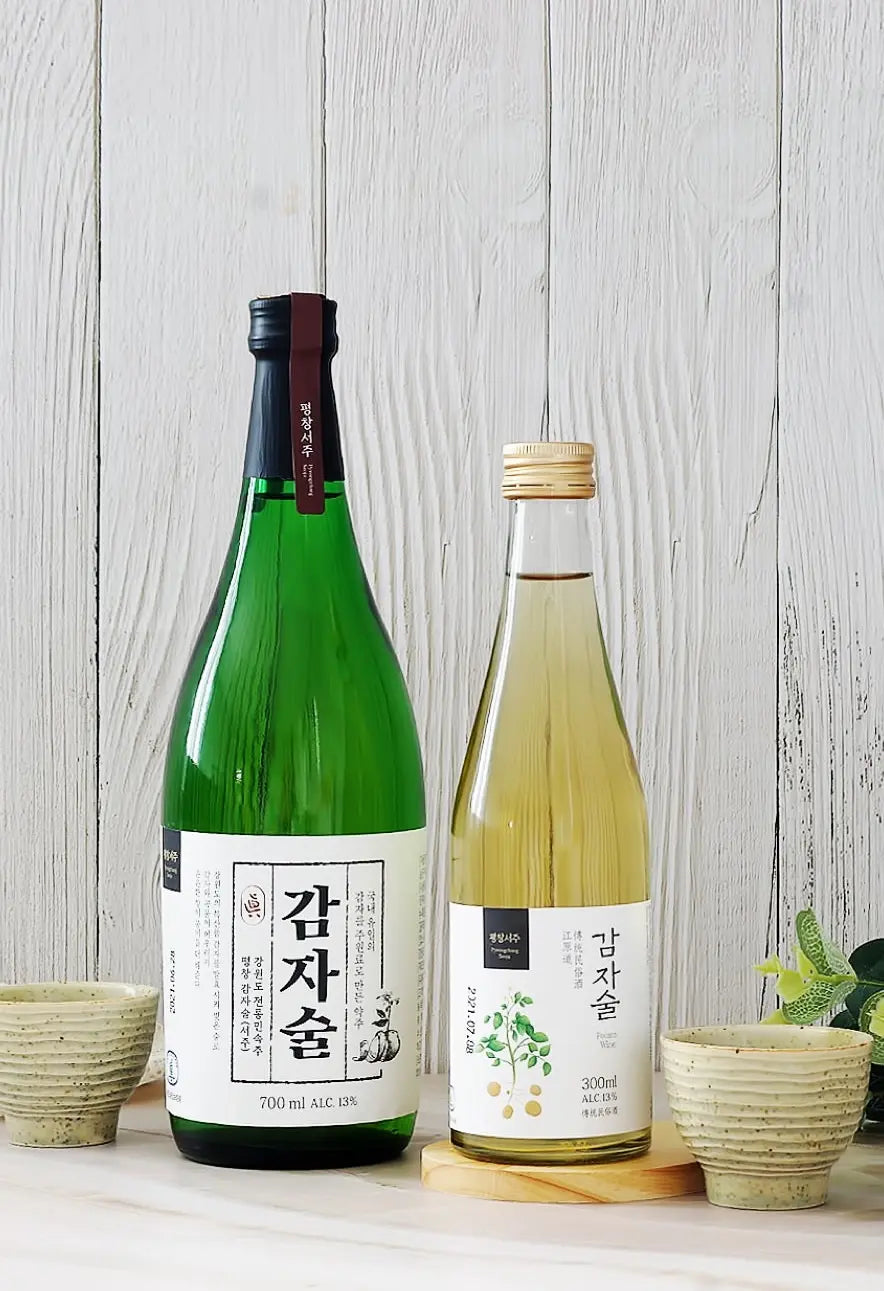 Two Korean rice wine bottles displayed on a light wooden surface against a white wood plank background, flanked by two small beige ceramic cups. Seoju Gamjasool - Odae Seoju - cult 