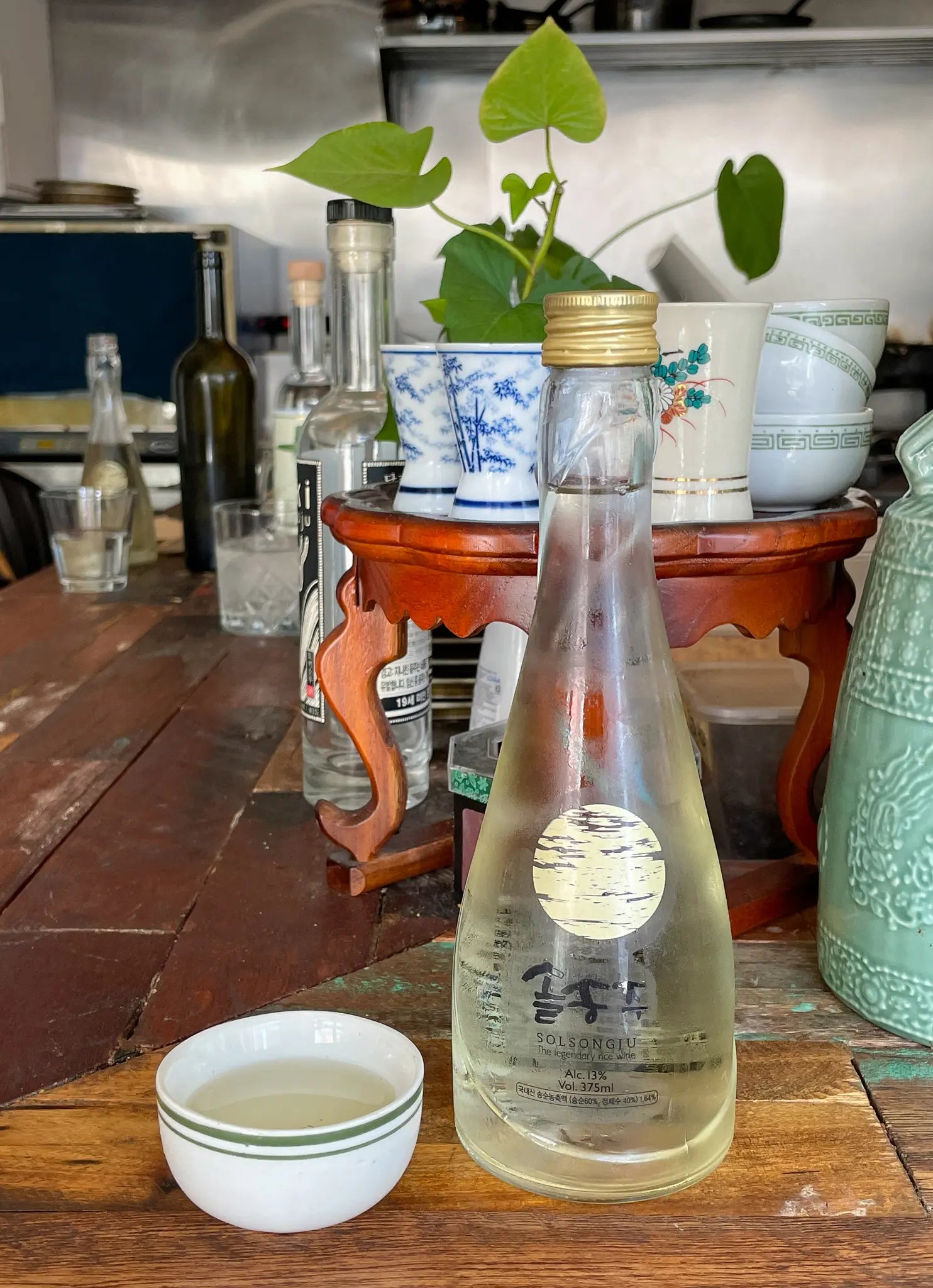 A clear glass bottle of Korean traditional liquor with a gold cap and a round gold label, placed on a rustic wooden table next to a small white ceramic cup. The background features a wooden stand with blue-and-white patterned ceramic cups, a green plant, and other bottles in a kitchen setting. Photo credit: drinkit @soolditor

Solsongju - pine yakju - cultsool