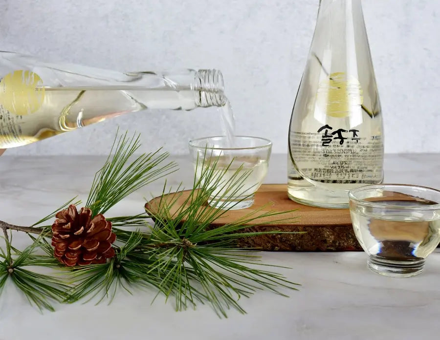 A clear glass bottle pouring pale golden liquid into a small glass cup, with another cup and a wooden slice on a light surface. Pine branches and a brown pinecone are arranged in the foreground, and the bottle has black Korean characters on the label.

Solsongju - pine yakju - cultsool