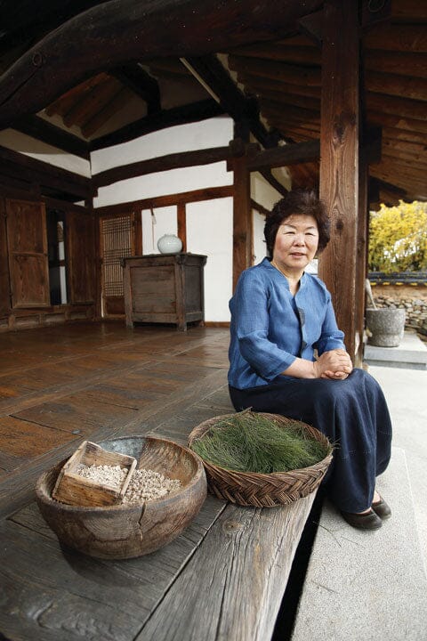 A woman — Korean Food Grand Master Park Hung-Sun — in a blue blouse and dark skirt sits on a wooden platform inside a traditional Korean house, with baskets of herbs and grains in the foreground.

Solsongju - pine yakju - cultsool