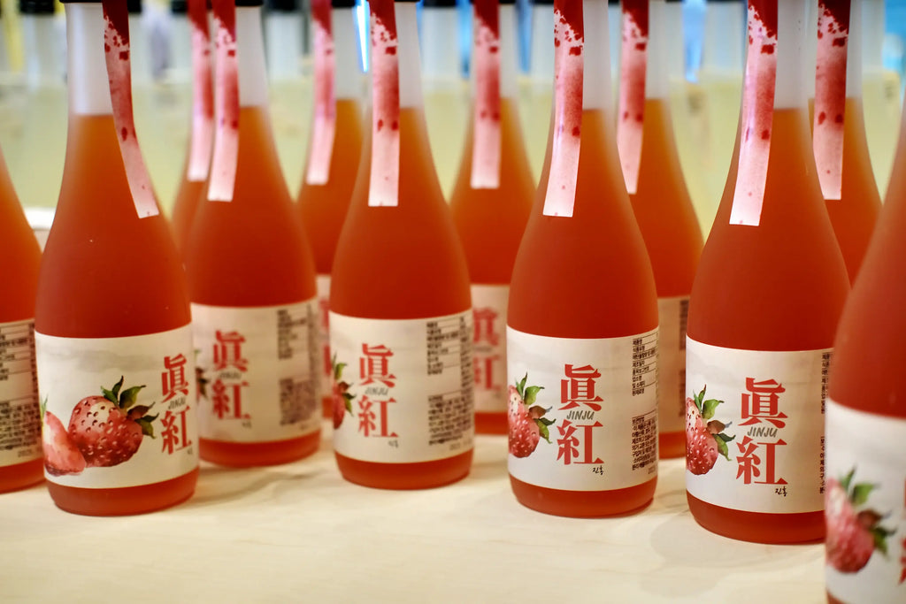 A row of Korean fruit wine bottles filled with a pinkish-red liquid, featuring white labels with red text and strawberry illustrations, set against a softly blurred background. Jinju Strawberry (Ddalgi) Wine - Good Tomorrow Brewery - cult.
