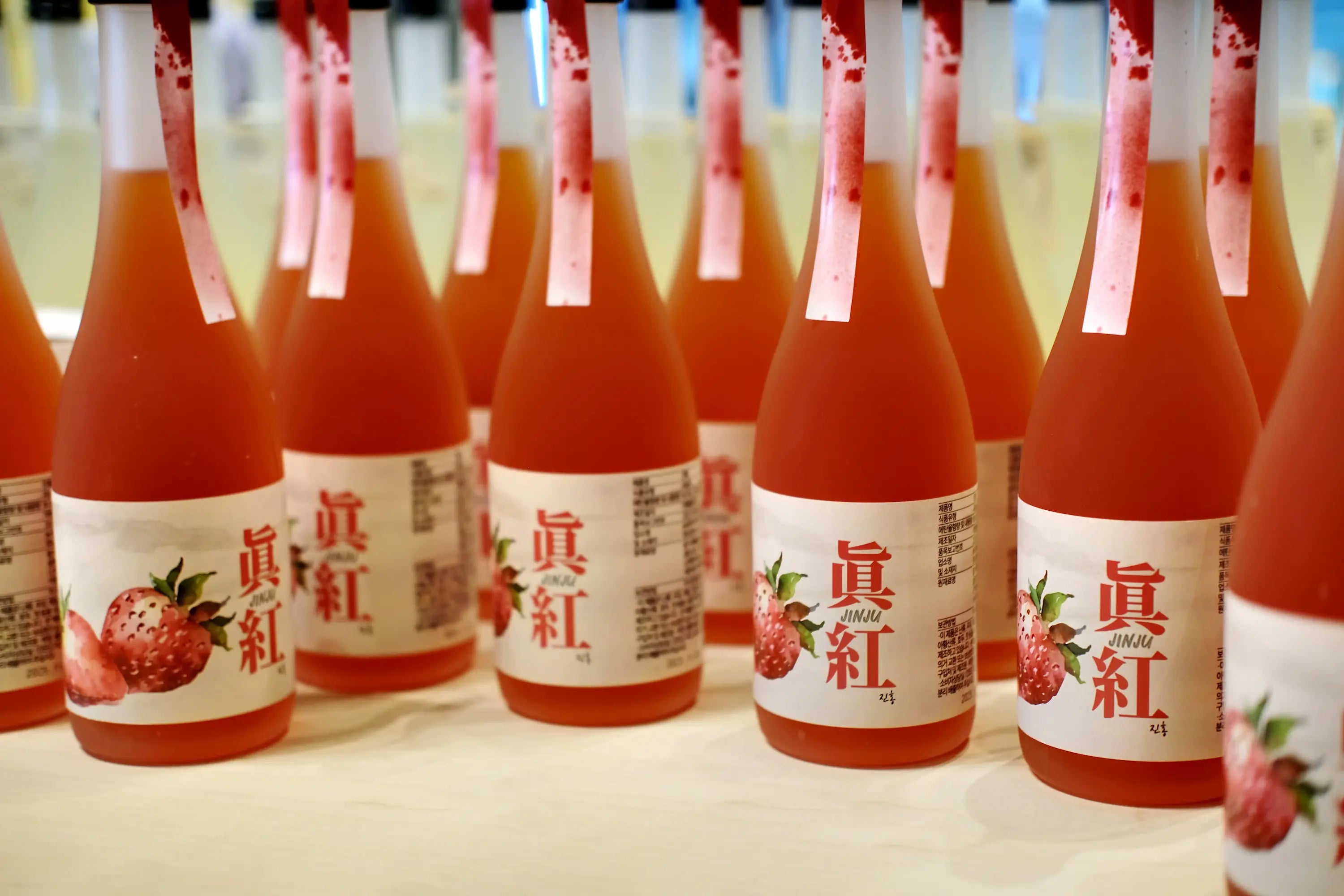 A row of Korean fruit wine bottles filled with a pinkish-red liquid, featuring white labels with red text and strawberry illustrations, set against a softly blurred background. Jinju Strawberry (Ddalgi) Wine - Good Tomorrow Brewery - cult.