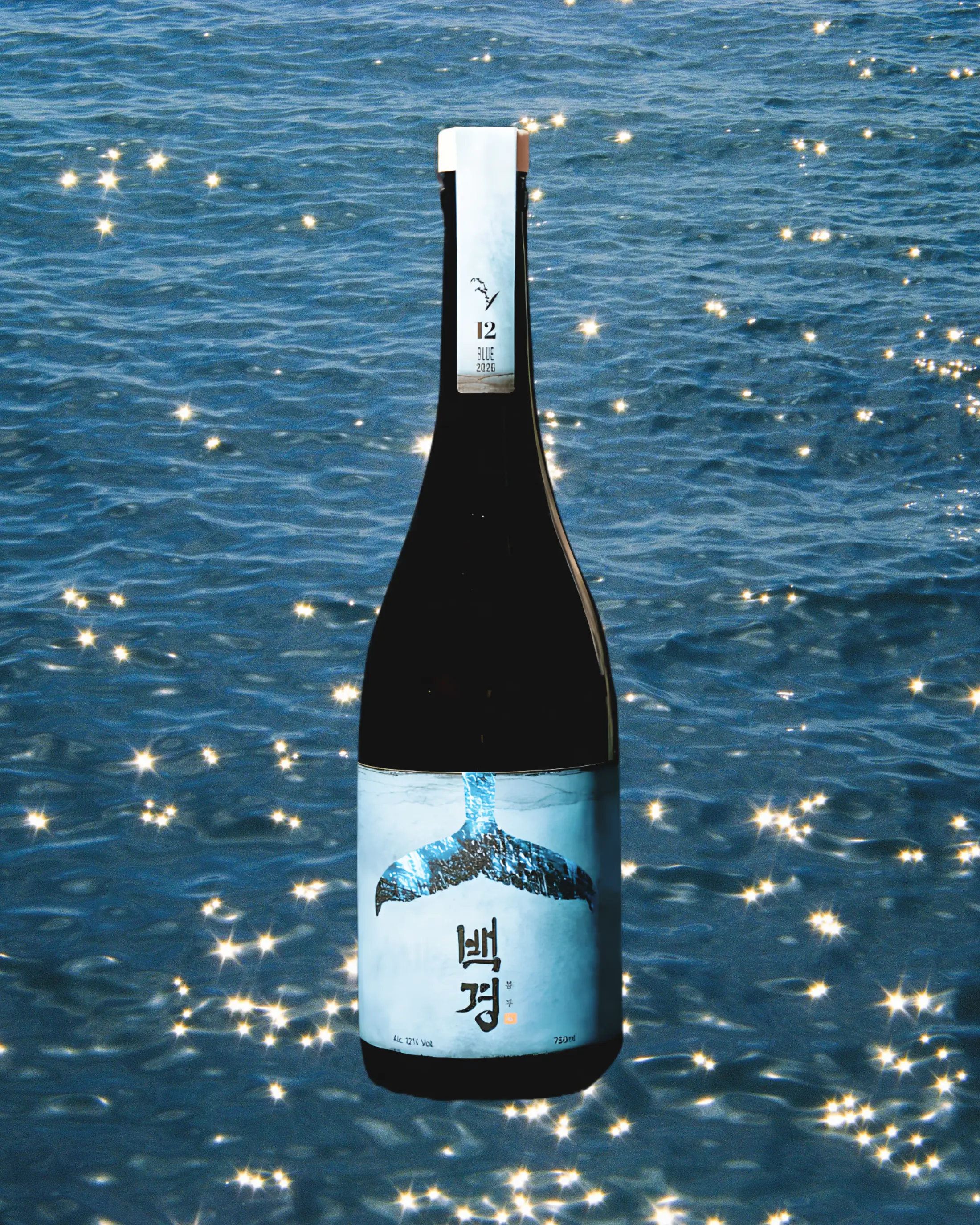 A dark glass bottle of Korean rice wine,  featuring a white label with a whale tail illustration and Korean text, set against a sparkling blue ocean background. Baekkyung Blue - yakju - cult
