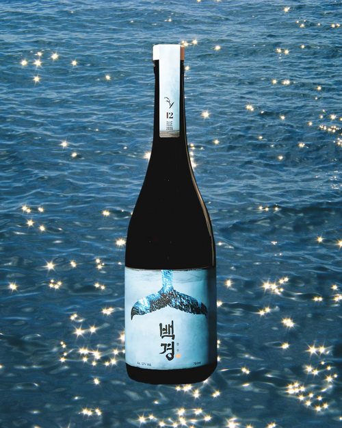 A dark glass bottle of Korean rice wine,  featuring a white label with a whale tail illustration and Korean text, set against a sparkling blue ocean background. Baekkyung Blue - yakju - cult