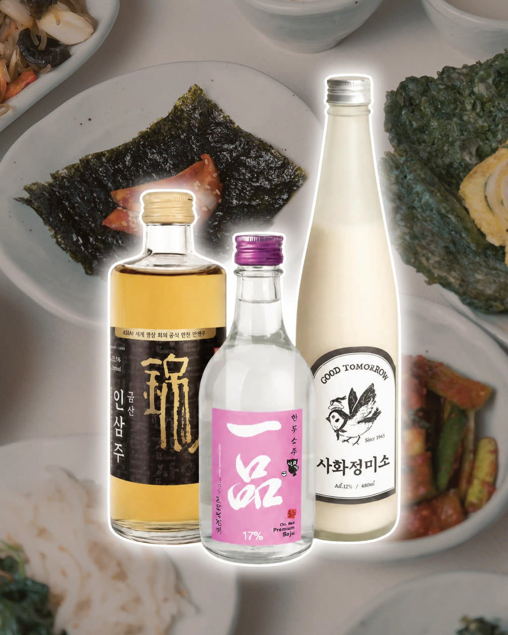 Three Korean beverage bottles centered over a blurred background of small Korean dishes like seaweed and cucumber kimchi. The left bottle contains a golden yellow liquid with a black label and gold text.