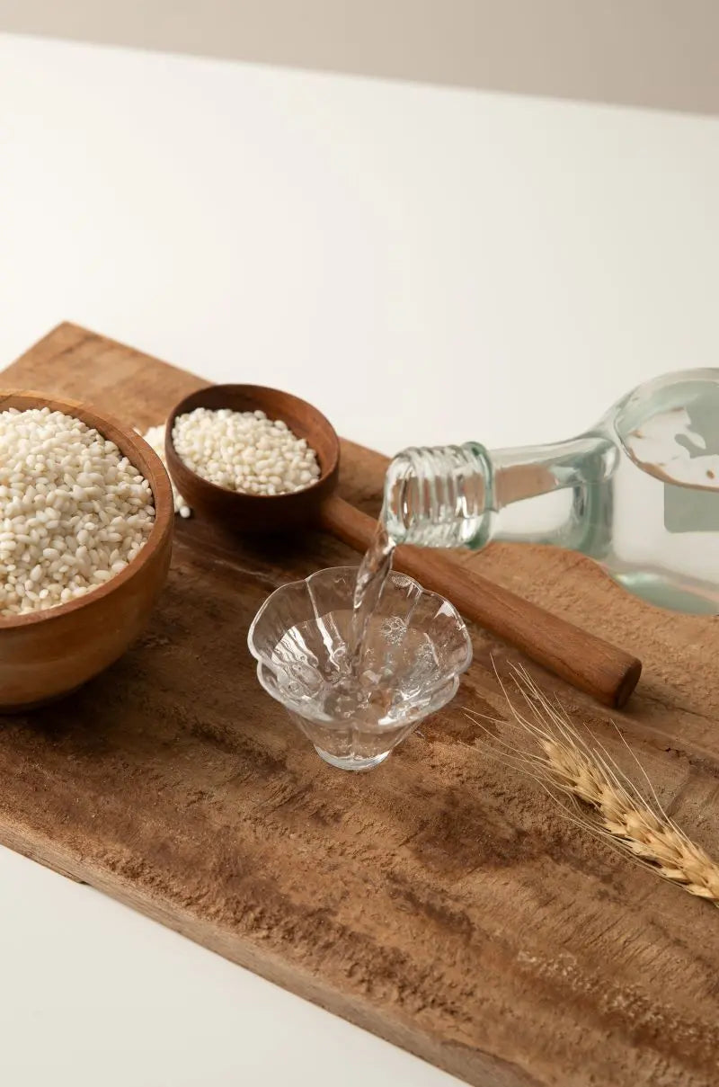 A clear glass bottle pouring soju into a small clear glass cup, arranged on a wooden board with bowls of rice and a rice stalk. Ilpoom Andong Soju 17% - distilled rice soju - cult 