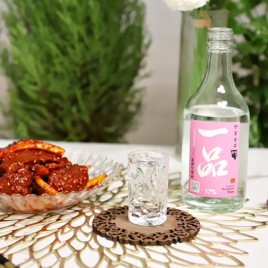 A clear glass bottle of Korean liquor with a pink label stands on a patterned table mat beside a small clear shot glass and a bowl of red braised food, with blurred greenery in the background. Ilpoom Andong Soju 17% - distilled rice soju - cult 