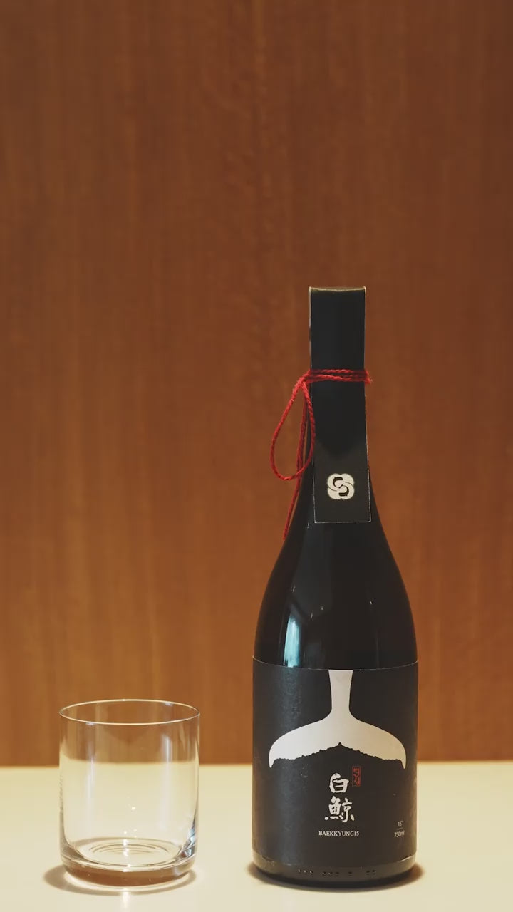 A close up of a black bottle of Korean rice wine. A person unties a red string around the neck. Golden liquid is poured into a glass. A man drinks the yakju. Baekkyung 15 Premium - cult