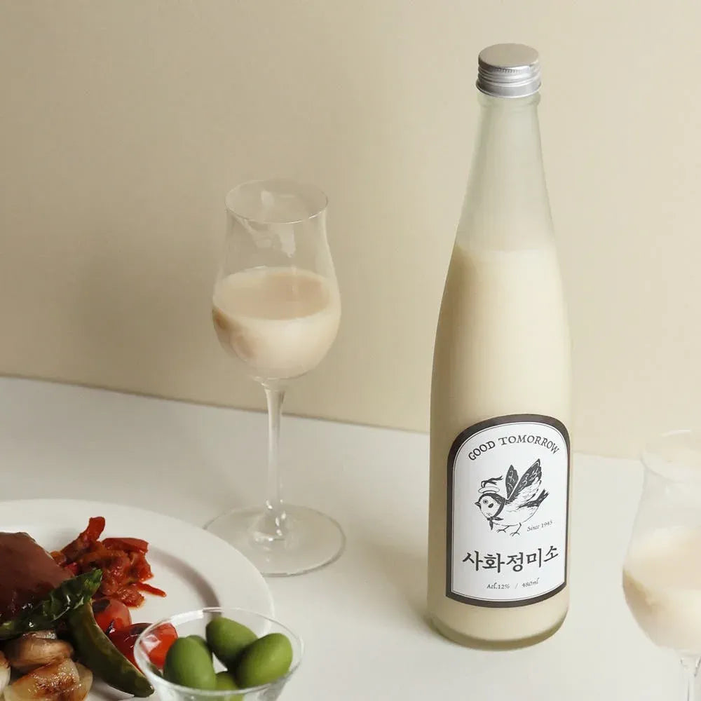 A tall, slender glass bottle of Korean makgeolli with a white label with black text and bird illustration stands on a white table beside two stemmed glasses filled with rice wine. A plate with food and green olives is partially visible on the left against a warm beige background.

Sahwajeongmiso Makgeolli - Good Tomorrow Brewery - 