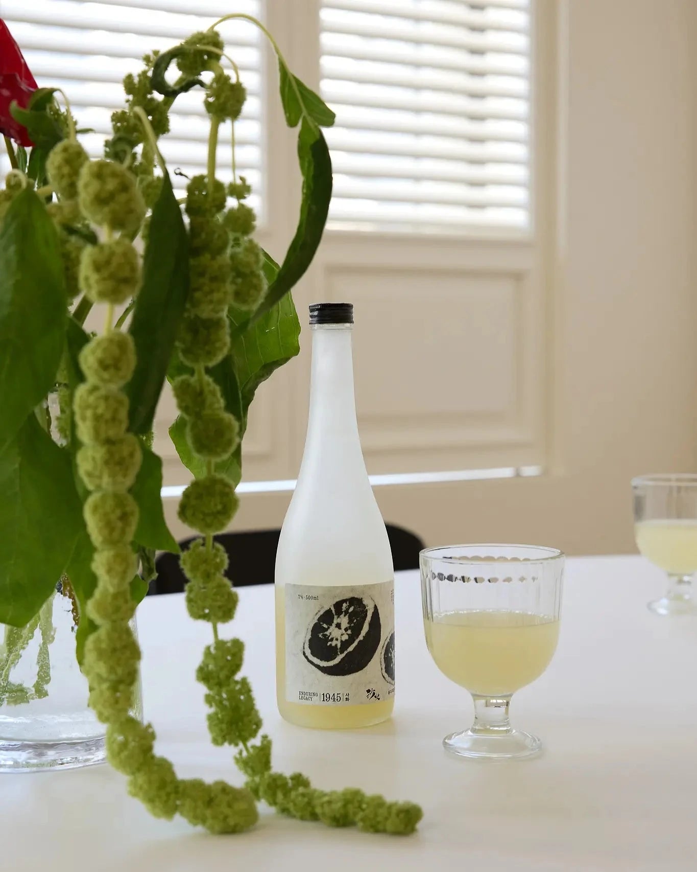 A frosted glass bottle of Korean citrus wine with a black cap and label, placed on a white table beside a two filled glasses, with green hanging flowers in the foreground and window blinds in the background.

Sahwa Yuzu - Good Tomorrow Brewery - cultsool