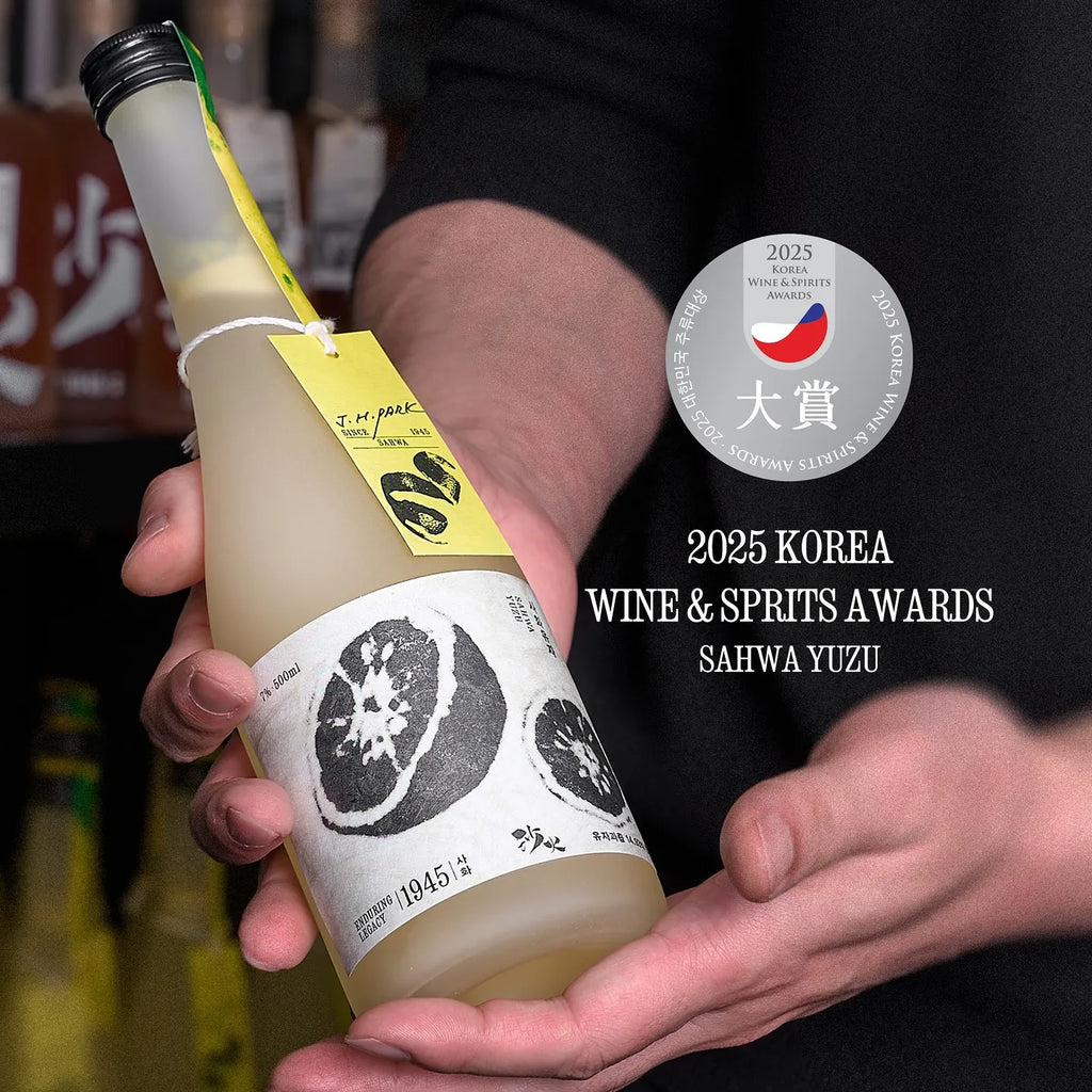 A person holds a bottle of Saha Yuzu Korean fruit wine in a dimly lit bar or restaurant setting. The bottle has a white label with black citrus graphics and a yellow hangtag. The words "2025 Korea Wine & Spirits Awards" and a silver badge graphic have been added.

Sahwa Yuzu - Good Tomorrow Brewery - cultsool