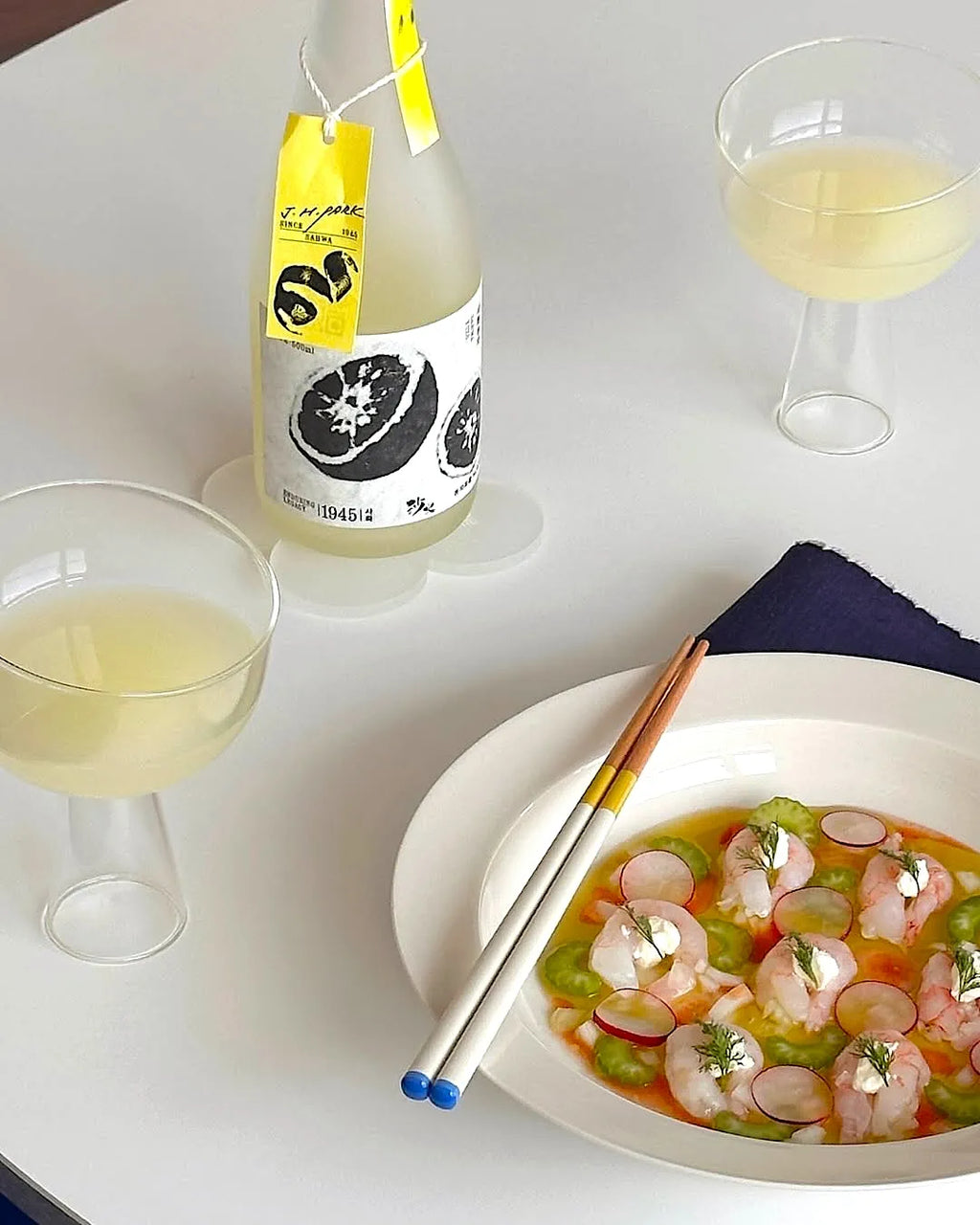 A bottle of cloudy citrus wine with a yellow tag and black-and-white label, styled on a white table with two glasses of pale yellow drink, a plate of shrimp ceviche and radish dish, and chopsticks.

Sahwa Yuzu - Good Tomorrow Brewery - cultsool