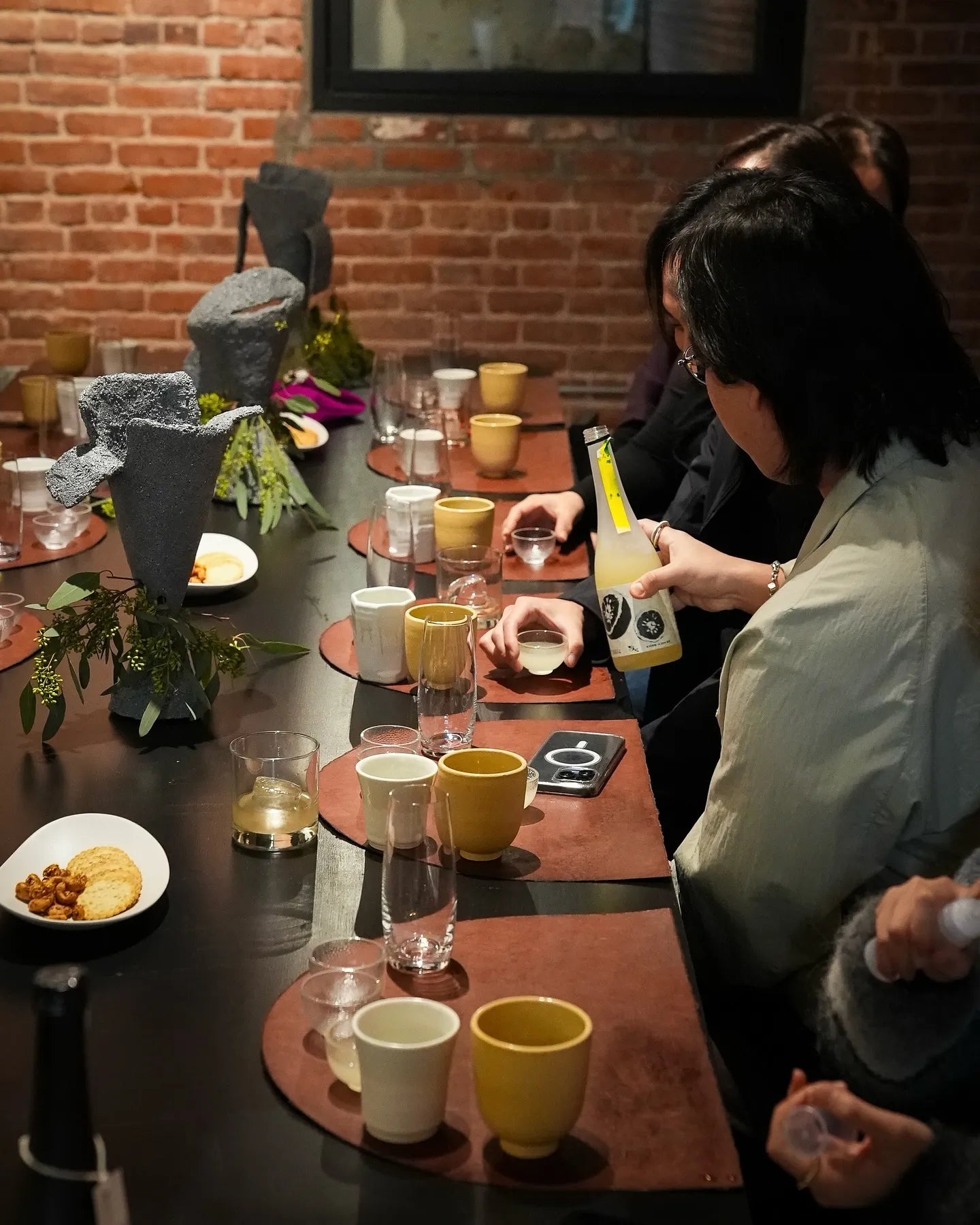 A group of people gathered around a dark wooden table in a restaurant or cafe setting, with a person pouring yellow citrus wine from a bottle into a glass. The table is decorated with ceramic cups in yellow, white, and brown, glassware, and small dishes, set against a rustic brick wall background.

Sahwa Yuzu - cultsool